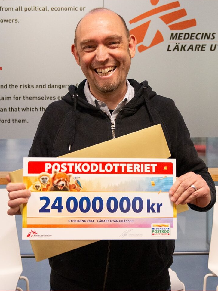 Generalsekreterare Oliver Schulz ler mot kameran med en stor check från Postkodlotteriet i händerna