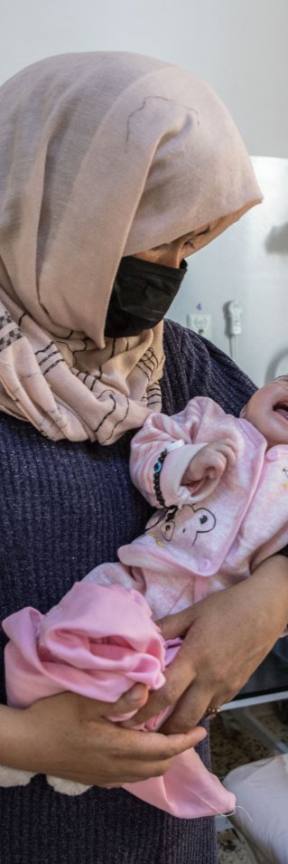 En kvinna med munskydd och slöja håller i en nyfödd bebis inne i en sjukhussal